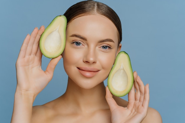 Pleased brunette young European female model holds halves of avocado near face