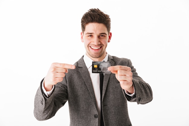 Pleased business man in jacket holding credit card and pointing on it  over white wall