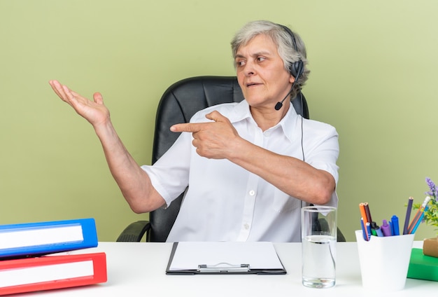 Pleased caucasian female call center operator on headphones sitting at desk with office tools pointing and looking at her empty hand