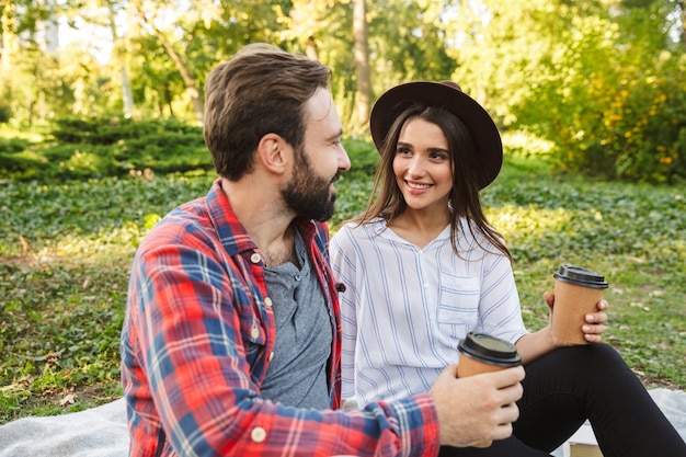 pleased couple man and woman dressed in casual wear drinking takeaway coffee while resting in green park