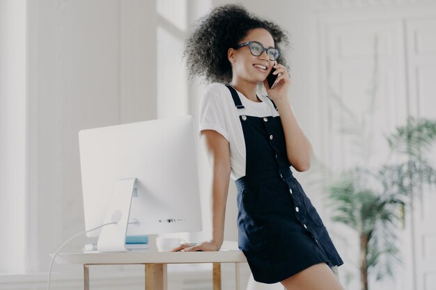 Pleased curly female office worker dressed in black sarafan leans at table poses in cabinet near computer monitor has telephone conversation discusses details of contract has smile on face