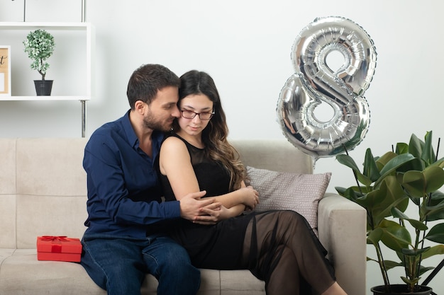 Pleased handsome man hugging and looking at pretty young woman in glasses sitting on couch in living room on march international women's day