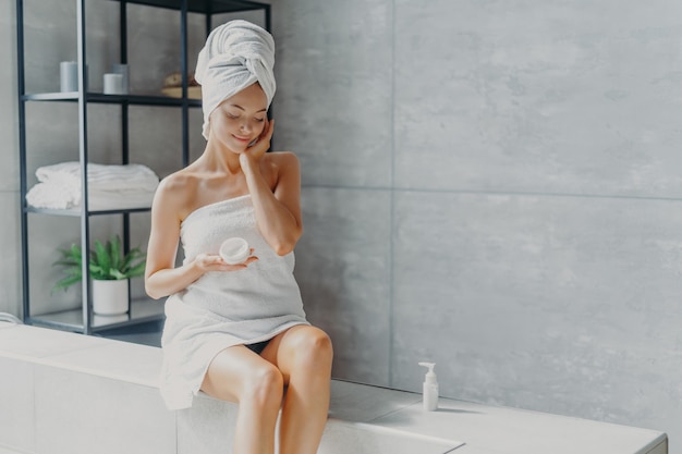 Pleased healthy Caucasian woman applies beauty cream on skin, poses in bathroom, undergoes hygienic treatments, cosmetic cream treatment, wrapped in towel, treats skin with hydrating product