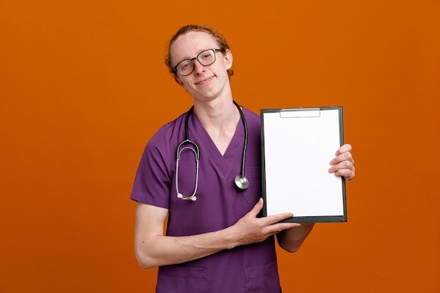 Pleased holding clipboard young male doctor wearing uniform with stethoscope isolated on orange background