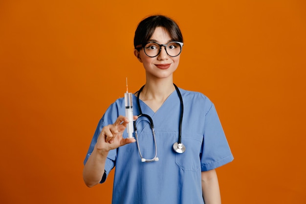 Pleased holding syringe young female doctor wearing uniform fith stethoscope isolated on orange background