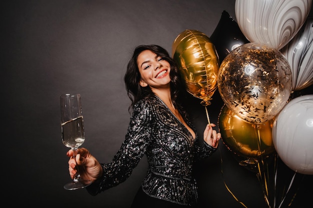 Pleased latin girl standing near party balloons Enchanting young woman drinking wine in her birthday