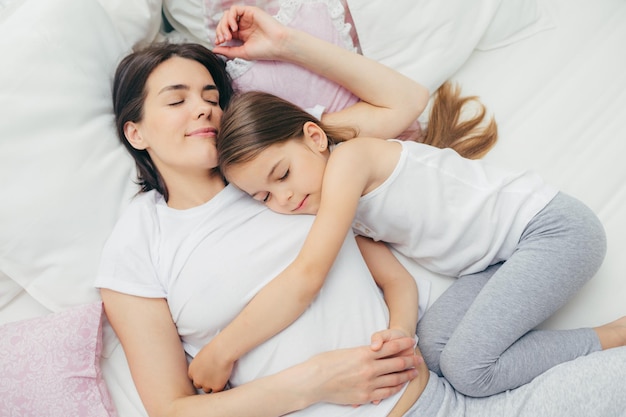 Pleased little child sleeps near her mother embraces with love has pleasant dreams lie on comfortable bed Mum and cute daughter have good sleep in bedroom Family sleeping and rest concept