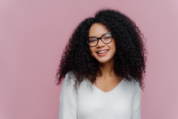 Pleased lovely woman with curly hairstyle, smiles gently at camera, wears optical glasses and white casual sweater