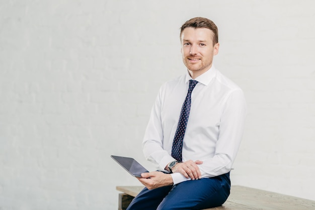 Pleased male entrepreneur makes payment online on digital tablet dressed in elegant white shirt tie and trousers sits on wooden table isolated over white background Business concept