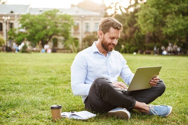 pleased man in business clothing, sitting on grass in park with legs crossed and working on silver laptop