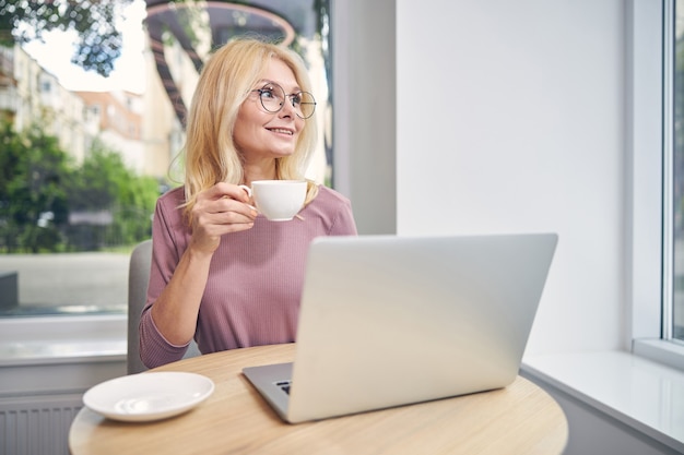 Pleased mature woman keeping smile on her face while sitting in front of her computer