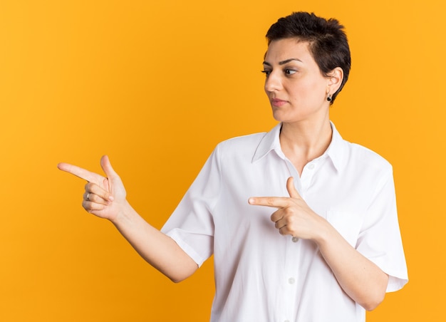 Pleased middle-aged woman looking and pointing at side isolated on orange wall