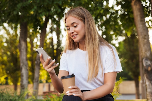 Pleased positive optimistic young lady sit in nature park using mobile phone drinking coffee.