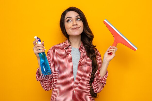 Pleased pretty caucasian cleaner woman holding squeegee and spray cleaner and looking up