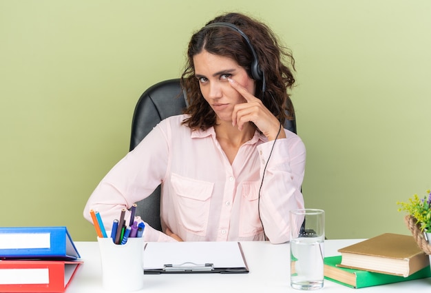 Pleased pretty caucasian female call center operator on headphones sitting at desk with office tools putting hand on her face isolated on green wall