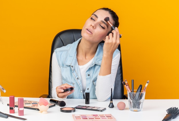 Pleased pretty caucasian woman sitting at table with makeup tools holding and applying blush with makeup brush isolated on orange wall with copy space