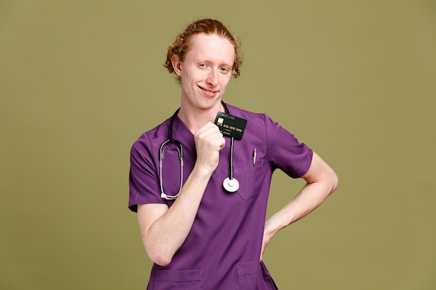 Pleased putting hand on hips young male doctor wearing uniform with stethoscope holding card isolated on green background