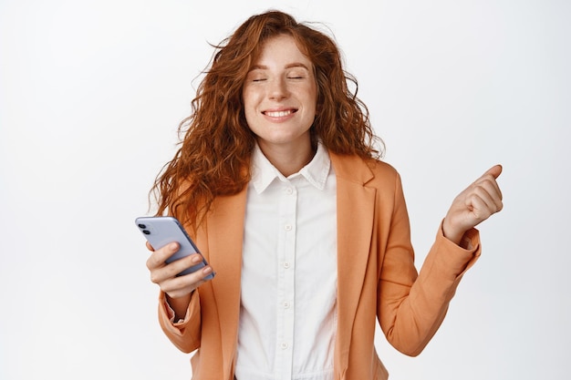 Pleased saleswoman holding her phone and triumphing smilling satisfied standing in suit against white background