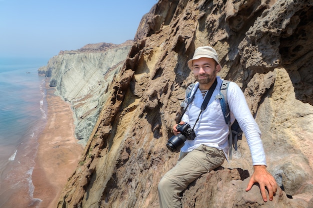 Pleased traveler with camera sits on edge of cliff, Hormuz Island, Hormozgan, Iran.