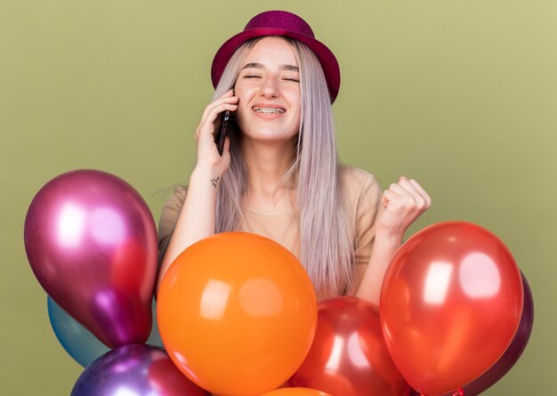 Pleased with closed eyes young beautiful girl wearing dental braces with party hat standing behind balloons speaks on phone 