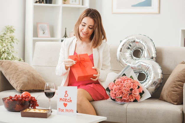 Pleased woman on happy women's day holding and looking at present sitting on sofa in living room