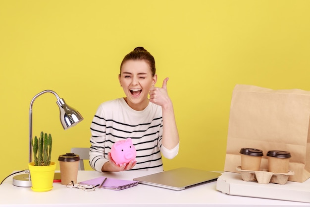 Pleased woman holding pig money box in hands saving showing thumb up winking sitting at workplace