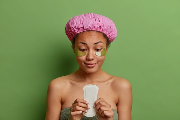 Pleased woman holds sanitary napkin applies collagen patches under eyes wrapped in bath towel wears waterproof hat isolated over green wall