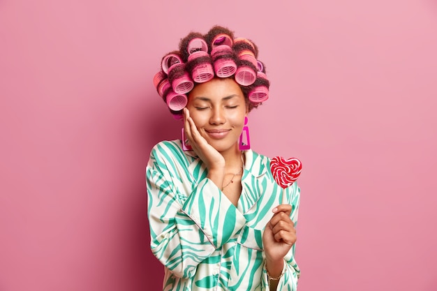 Pleased woman keeps eyes closed enjoys domestic atmosphere makes hairstyle applies hair rollers holds tasty candy wears dressing gown isolatd over pink wall