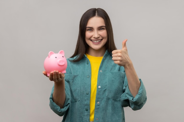 Pleased woman with dark hair holding pig money box in hands saving showing thumb up