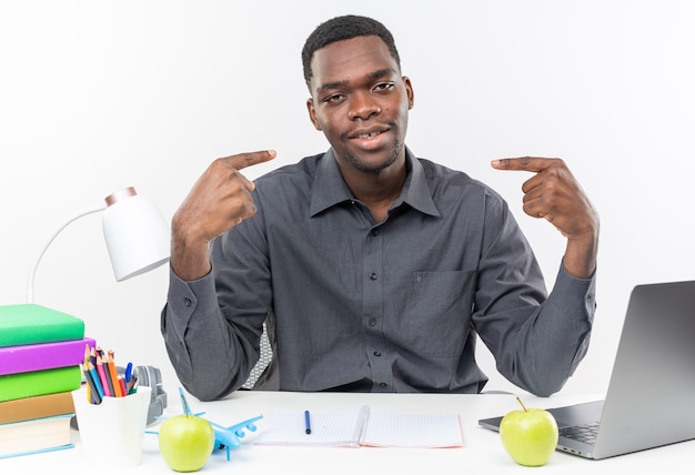 Pleased young afro-american student sitting at desk with school tools pointing at himself 