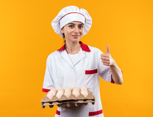 Pleased young beautiful girl in chef uniform holding batch of eggs showing thumb up isolated on orange wall