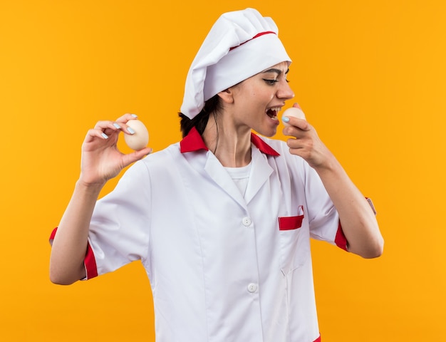 Pleased young beautiful girl in chef uniform holding eggs and trying it isolated on orange wall