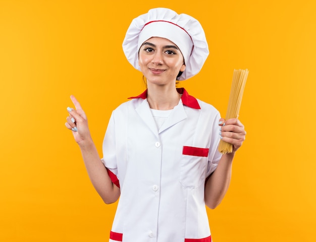 Pleased young beautiful girl in chef uniform holding spaghetti points at side 