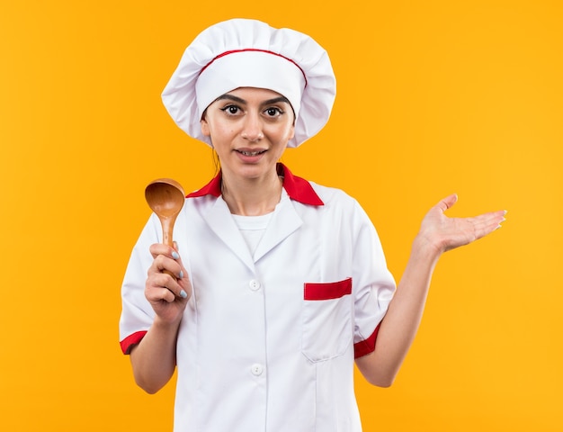 Pleased young beautiful girl in chef uniform holding spoon spreading hand 