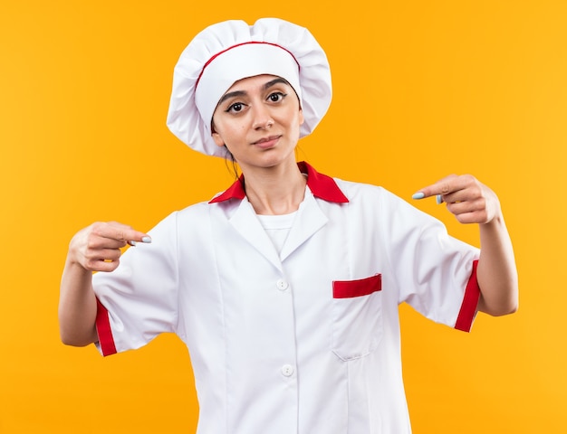 Pleased young beautiful girl in chef uniform points at herself isolated on orange wall