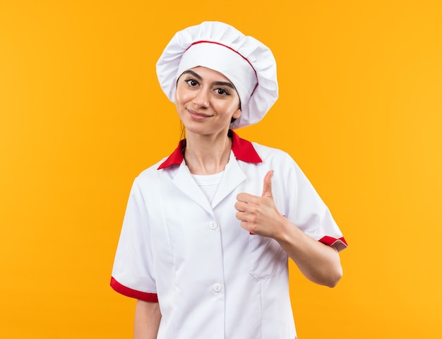 Pleased young beautiful girl in chef uniform showing thumb up isolated on orange wall