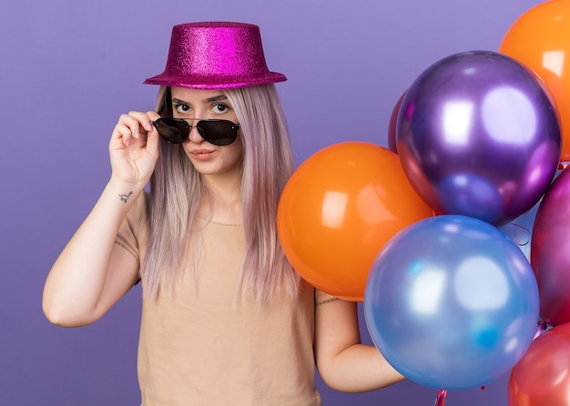 Pleased young beautiful girl wearing party hat with glasses holding balloons 