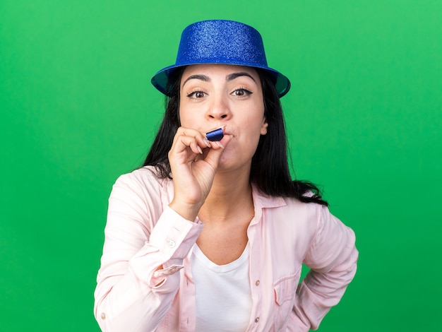 Pleased young beautiful woman wearing party hat blowing party whistle putting hand on hip