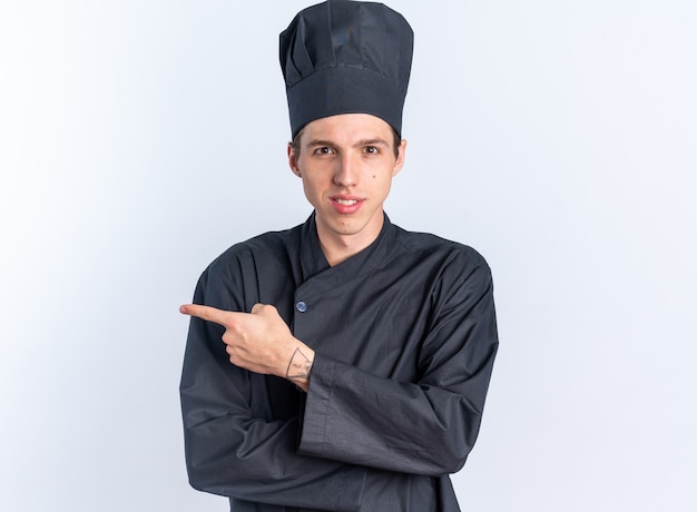 Pleased young blonde male cook in chef uniform and cap looking at camera pointing at side isolated on white wall with copy space