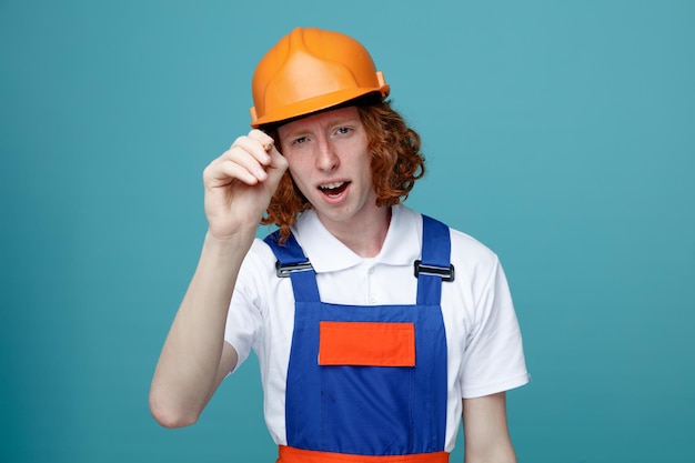 Pleased young builder man in uniform holding out marker pen to camera isolated on blue background