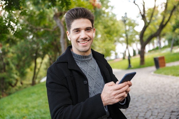 pleased young cheerful man in casual clothing walking outdoors in green park using mobile phone listening music with earphones.