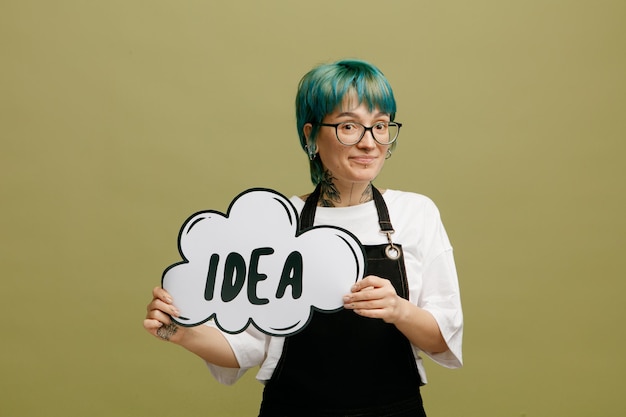 Pleased young female barber wearing glasses uniform looking at camera showing idea bubble isolated on olive green background