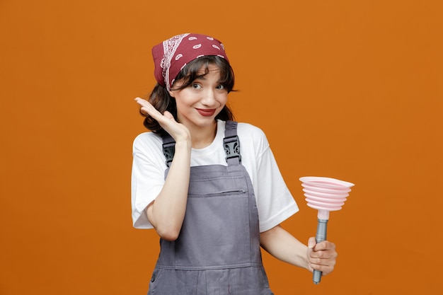 Pleased young female cleaner wearing uniform and bandana holding plunger looking at camera showing empty hand isolated on orange background