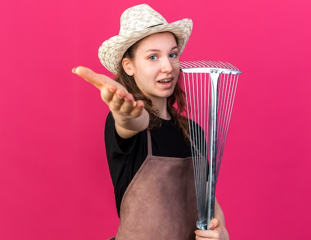 Pleased young female gardener wearing gardening hat holding leaf rake and holding out hand at camera 