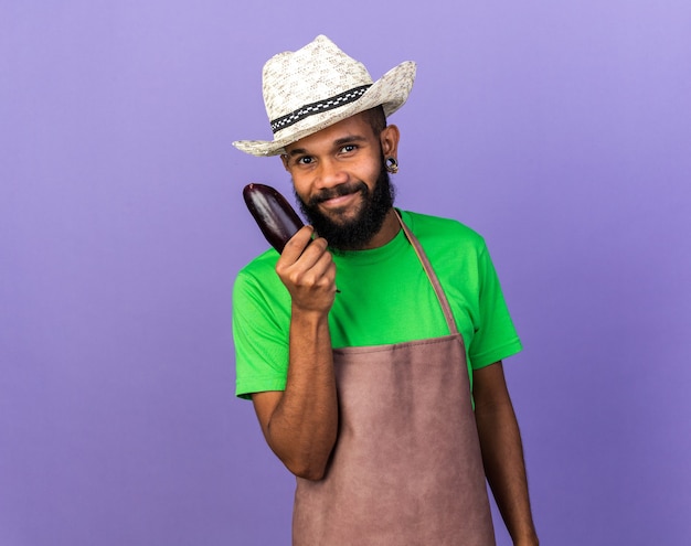 Pleased young gardener afro-american guy wearing gardening hat holding eggplant 