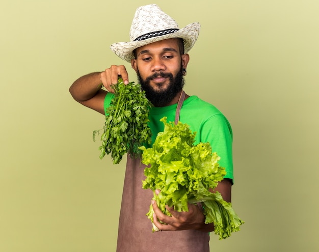 Pleased young gardener afro-american guy wearing gardening hat holding out salad at camera 