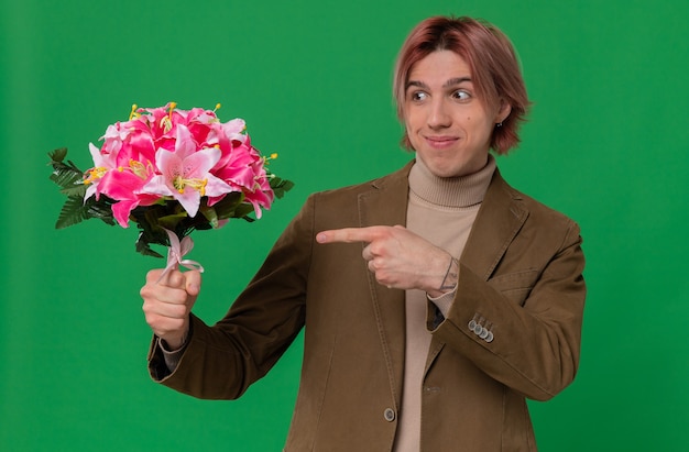 Pleased young handsome man holding and pointing at bouquet of flowers