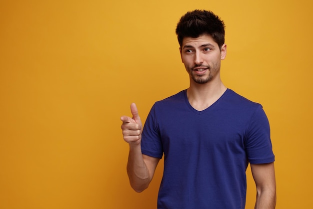 Pleased young handsome man looking and pointing at side on yellow background with copy space