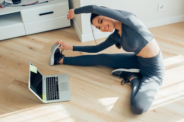 Pleased young lady in sport clothes stretching legs in front of a laptop and smiling while bending towards the foot