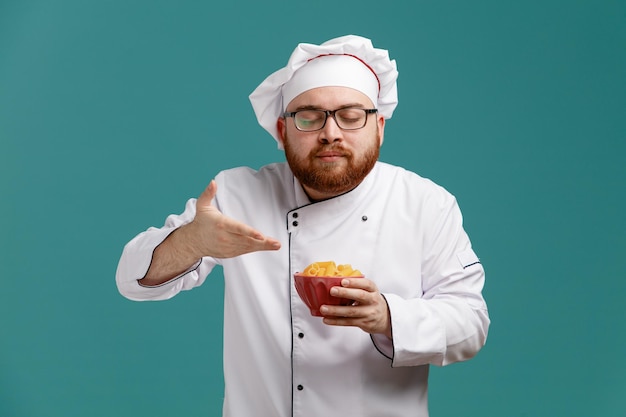 Pleased young male chef wearing glasses uniform and cap holding bowl of macaronis pointing with hand at it smelling aromas of food with closed eyes isolated on blue background